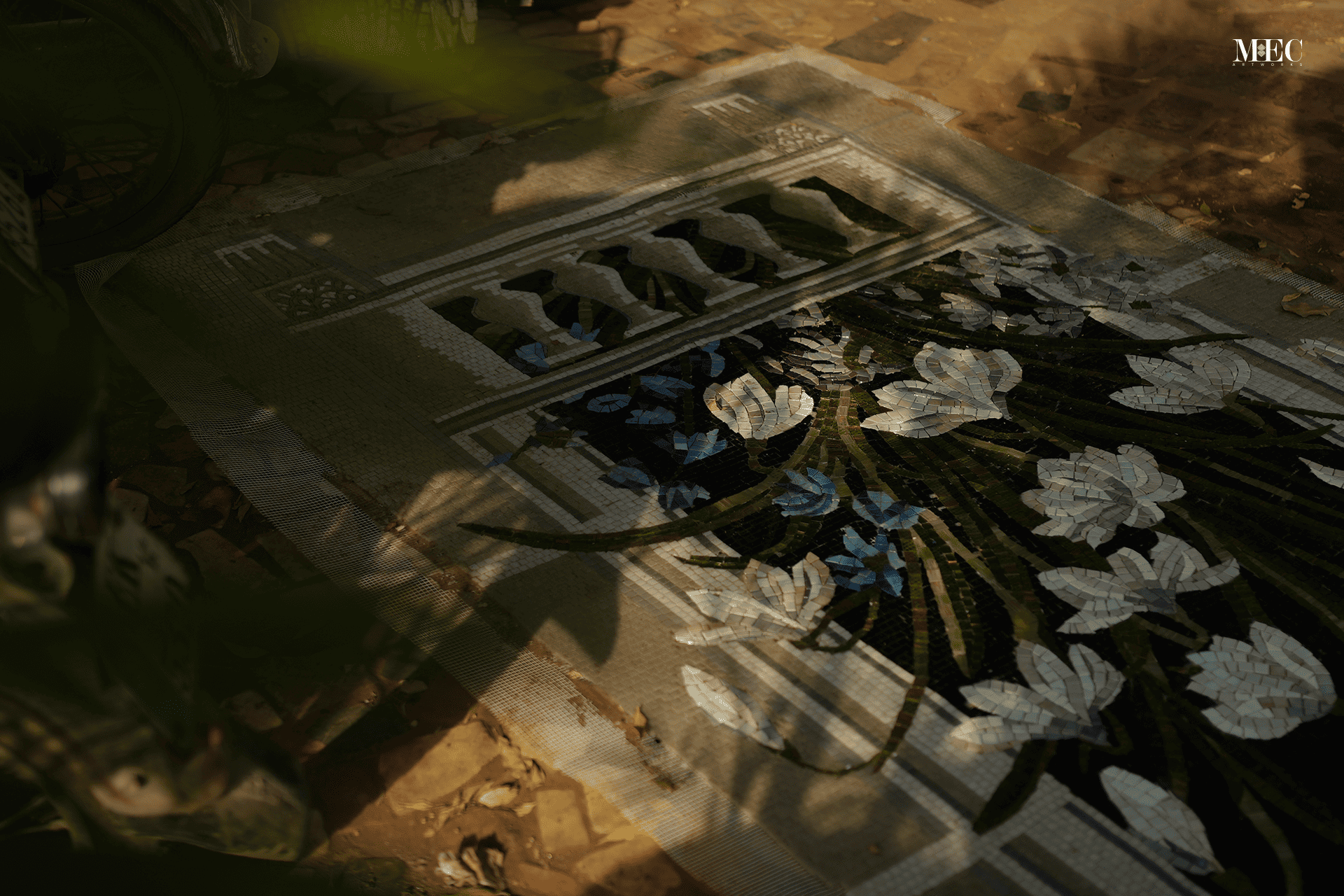 A detailed mosaic artwork of white flowers and green leaves is partially completed on a fabric canvas, laid on the ground amidst fallen leaves and dappled sunlight.
