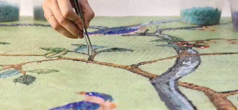 Artist placing mosaic tiles with tweezers to complete a colorful nature scene on green background.
