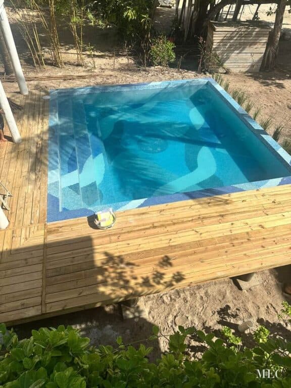 Small outdoor pool with abstract blue mosaic tiles, wooden deck, and tropical landscape background.