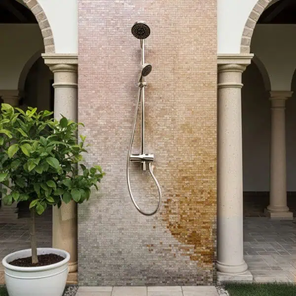 Terracotta Glow Outdoor Shower Mosaic Outdoor shower with stainless steel fixtures mounted on a mosaic wall featuring a gradient of beige to rose-gold tiles, framed by stone columns and a potted green plant on a tiled and pebbled base.