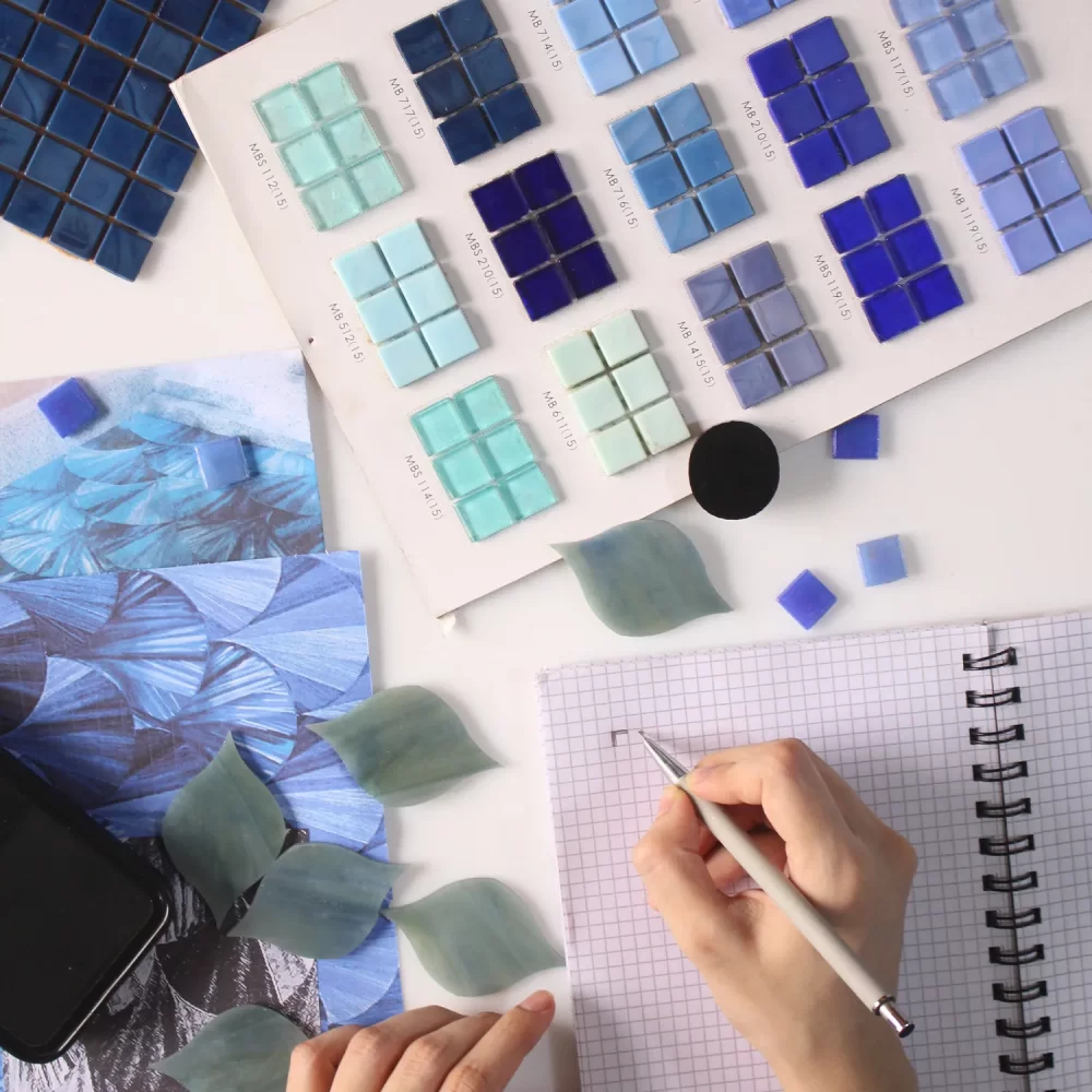 About Hand designing tile layout with blue mosaic samples, color swatches, leaves, and a gridded sketchbook.