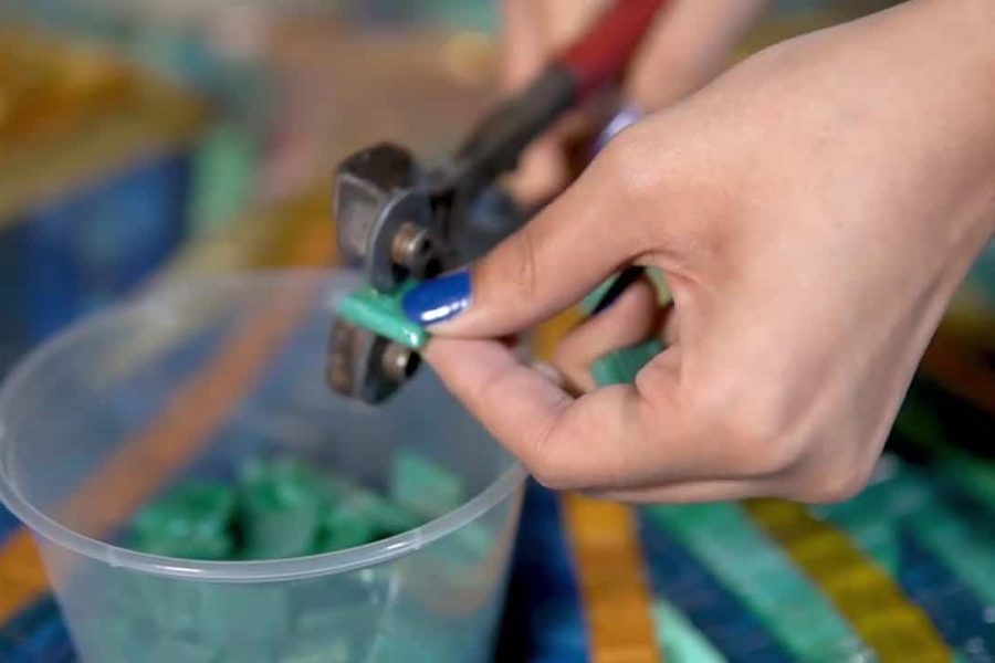 Close-up of hand using nippers to cut green mosaic tile above plastic container on table.
